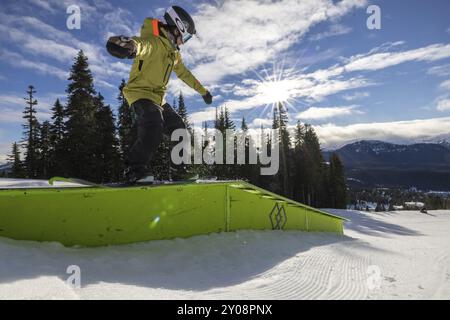 Ein Snowboarder führt einen Trick auf einer hellgrünen Schiene mit einer wunderschönen Schneelandschaft und Sonnenlicht im Hintergrund aus Stockfoto