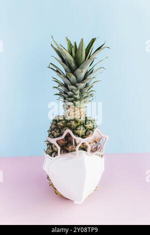 Sicheres Reise- und Urlaubskonzept mit Ananas in Sonnenbrille und schützender Gesichtsmaske. Rosa Gläser in Form eines Sterns. Entspannung am Meer. Rosa Stockfoto