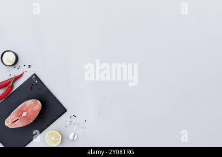 Grauer Betonhintergrund flach mit Schnitt aus rotem rohem Lachs Fischfiletsteak auf schwarzem Schneidebrett, Zitrone, Pfeffer würzen, Salz, Mayonnaise-Sauce Stockfoto