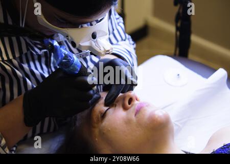 Eine Kosmetikerin trägt schwarze Handschuhe und eine Lupe und schminkt die Augenbrauen einer Frau in einem Schönheitssalon dauerhaft Stockfoto