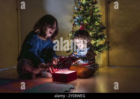 Ein kleiner Junge und ein Mädchen sitzen auf dem Boden vor einem Weihnachtsbaum und öffnen ein rotes Geschenk Stockfoto