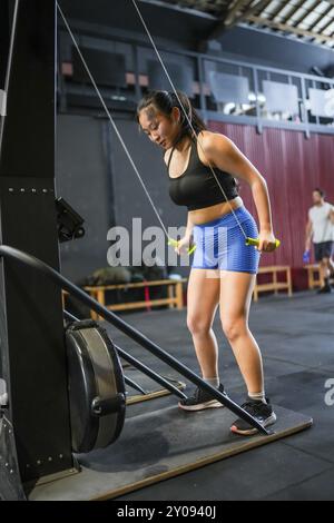 Vertikales Foto in voller Länge von einer asiatischen Frau, die in einem Fitnessstudio mit einem Pulldown-Lat-Gerät trainiert Stockfoto
