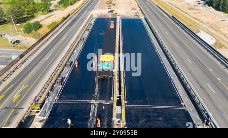 Luftaufnahmen einer Baustelle auf einer Autobahn mit großen Asphaltverlegemaschinen und Arbeitern. Die Website zeigt eine teilweise fertiggestellte bridg Stockfoto