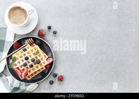 Stapel von zuckerpulverförmigen Waffeln mit Honig auf einem Teller auf dem Handtuch und grauen Beton- oder Schiefertisch mit Heidelbeere, gehackten Erdbeeren und Minzblättern Stockfoto