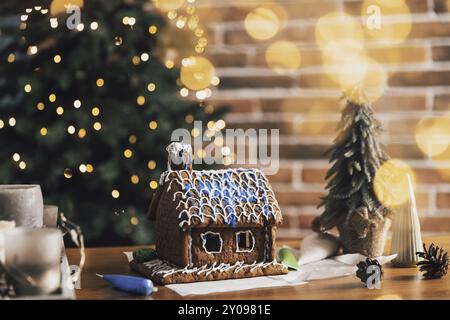 Kleines hausgemachtes Lebkuchenhaus auf einem Tisch mit Dekorationen, Kerzen im Wohnzimmer dekoriert mit unscharfen Neujahrslichtern, Weihnachtstanne Stockfoto