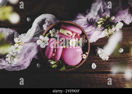 Blick von oben auf rosa Erdbeermakaron-Kekse in Keramikschüssel auf braunem Holz-Terrassendielen, weiße Apfelblüten. Farbenfrohe Komposition Stockfoto