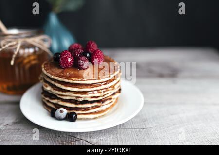 Saftige Pfannkuchen mit Beeren und Honig auf einem weißen Teller, Löffel, Glas, Holztisch. Stockfoto