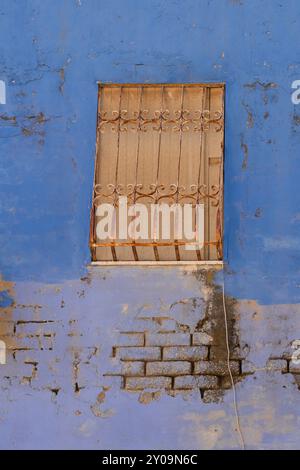 Ein Fenster in einem alten Gebäude, blockiert mit Holzbrettern und rostigen Sicherheitsbügeln. Die blaue und lila Farbe an der Gebäudewand löst sich ab, es liegt vor Stockfoto