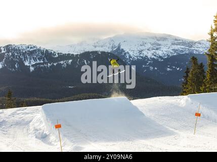 Snowboarder mitten im Sprung auf einem verschneiten Berghang mit immergrünen Bäumen und klarem Himmel Stockfoto