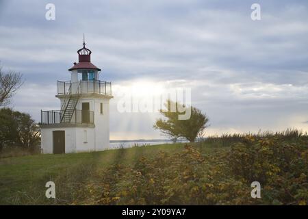 Leuchtturm, Spodsbjerg Fyr in Huntsted an der Küste Dänemarks. Die Sonne scheint durch die Wolken. Wiese mit Bäumen. Landschaftsfoto vom Meer Stockfoto