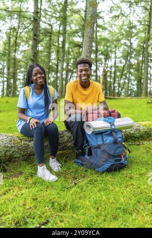 Vertikales Porträt zweier junger männlicher und weiblicher afrikanischer Wanderer, die auf einem lächelnden Baumstamm ruhen, umgeben von grünen Bäumen Stockfoto