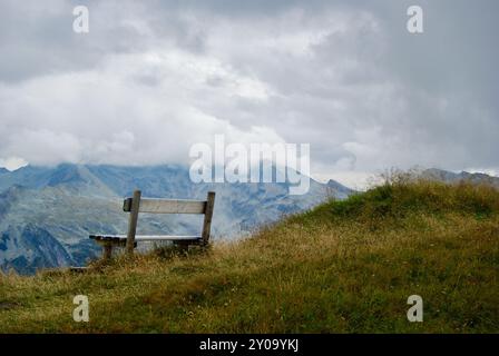 Nahaufnahme einer Holzbank mit Blick auf eine atemberaubende Berglandschaft an einem trüben Tag mit grauen Wolken und Nebel über den Gipfeln im Herbst in Österreich. Stockfoto