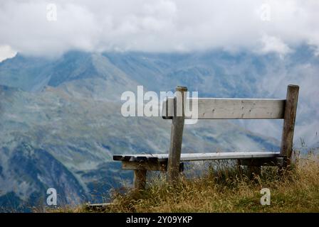 Nahaufnahme einer Holzbank mit Blick auf eine atemberaubende Berglandschaft an einem trüben Tag mit grauen Wolken und Nebel über den Gipfeln im Herbst in Österreich. Stockfoto