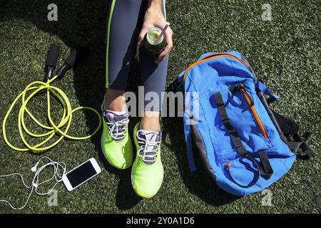 Sportliche Frau mit grüner Saft sitzen auf Rasen mit Springseil, Handy und Rucksack Stockfoto