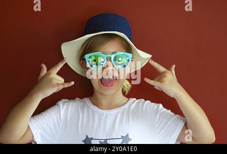 Ziemlich emothional Kind trägt einen Hut und eine Sonnenbrille auf einem roten Hintergrund. Platz kopieren, Tageslicht Stockfoto