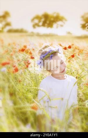 Kleines, fröhliches Mädchen mit einem selbstgewebten Kornblumenkranz am Mittsommer in Schweden, am Rande eines Kornfeldes mit Mohn und Kornblumen. Stockfoto