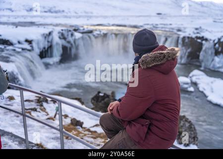 Ein Wandermann sitzt auf dem gefrorenen Godafoss Wasserfall bei Sonnenuntergang im Winter, Island, Europa Stockfoto