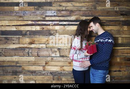 Ein junges Paar in der Nähe einer Holzwand feiert Weihnachten mit Geschenk. Liebes- und Beziehungskonzept Stockfoto