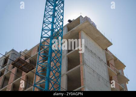 Ein blauer Kran hebt Betonblöcke an die Spitze eines im Bau befindlichen Wohngebäudes. Stockfoto