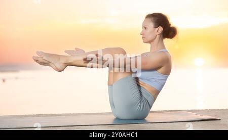 Fleißige junge Frau Trainerin, die Yoga-Pose auf dem Boot übt, Ubhai Padangushthasan auf Matte am Meer bei Sonnenuntergang. Philosophie des aktiven Lebensstils Stockfoto