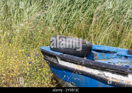 Das alte blaue Fischerboot liegt vergessen im Unkraut, ein Reifen liegt auf der Seite, ein Symbol der Vernachlässigung und des Vergehens der Zeit Stockfoto