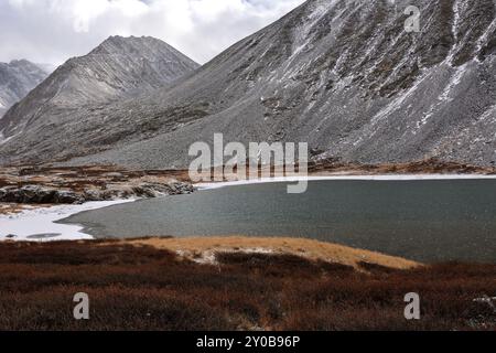 Bewachsen mit rotem Moos, die Ufer eines großen Sees mit Eis an den Rändern am Fuße eines hohen Berges, der mit dem ersten Schnee auf einer bewölkten Fläche bestreut ist Stockfoto