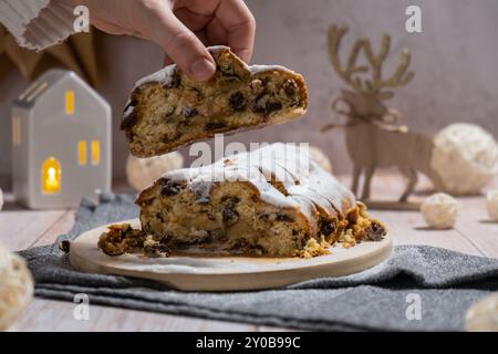 Weibliche Hand nimmt ein Stück Weihnachtsstollen in Weihnachtsbeleuchtung und Dekoration. Traditionelles süßes Obstbrot mit Puderzucker festliches Gebäck Dessert. Holiday Baking Food Konzept. Stockfoto