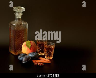 Pfirsichlikör mit Zimt und Anis, hausgemachter Alkohol in Kristallgläsern, Gewürze und reife Früchte auf schwarzem Hintergrund. Nahaufnahme. Stockfoto