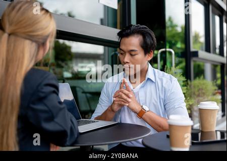 Ein seriöser, fokussierter asiatischer Mann hat ein informelles Treffen mit seinem Versicherungsagenten in einem Café und diskutiert, während er auf einen Laptop-Bildschirm schaut Stockfoto