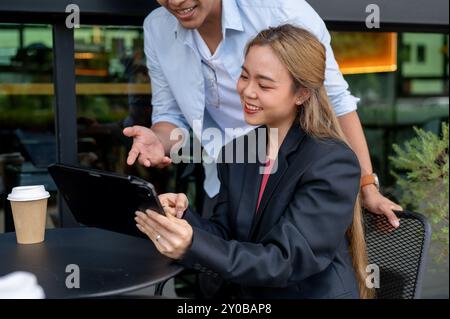 Zwei positive asiatische Kollegen, eine Frau und ein Mann, diskutieren über Arbeit und tauschen Ideen aus, während sie sich während einer informellen Sitzung einen Tablet-Bildschirm ansehen Stockfoto