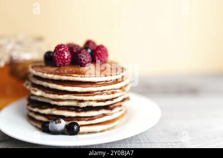 Saftige Pfannkuchen mit Beeren und Honig auf einem weißen Teller, Löffel, Glas, Holztisch. Stockfoto