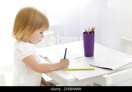 Seitenansicht des blonden, haarigen kleinen Mädchens, das am Tisch sitzt und zu Hause in Notizbuch mit bunten Bleistiften schreibt Stockfoto