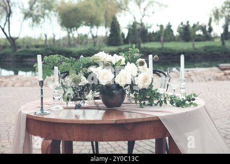 Kerzen und Vase mit weißen Rosen auf einem runden Tisch in der Nähe von Weingläsern während eines romantischen Datums im Garten Stockfoto