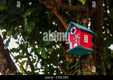 Ein rotes Vogelhaus in einem Baum. Das Vogelhaus hat einen Rettungsring, Sternfische, Möwen und Leuchtturm darauf. Stockfoto