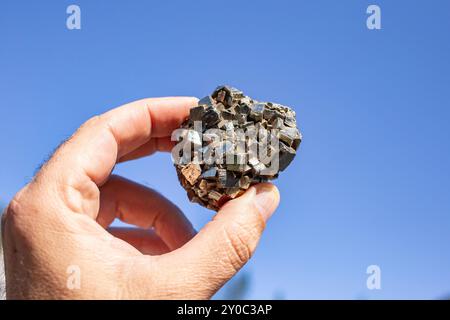 Männliche Hand, die einen goldenen Mineral-Pyrit-Stein vor blauem Himmel hält, im Sonnenlicht töricht Gold Stockfoto