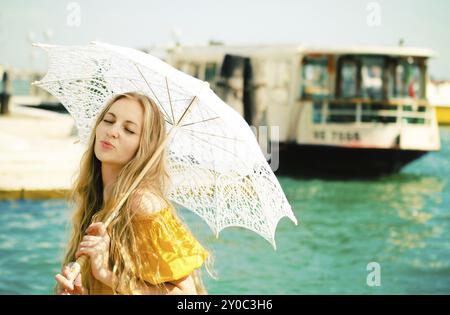 Junge glückliche Frau in gelbem Kleid, die einen Schirm auf dem Meeresboothintergrund hält Stockfoto