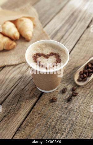 Papierbecher Kaffee mit Herzform, Creme, Schaum, Croissants bündeln sich am Sacktuch, getrocknete geröstete Kaffeebohnen im Löffel am alten Holztisch Stockfoto