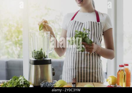 Junge lächelnde Frau, die in der Küche mit einem elektronischen Mixer grünen Frühlings-Smoothie kocht. Nahaufnahme von schönen, glücklichen Mädchen, die Hände legen Blätter i Stockfoto