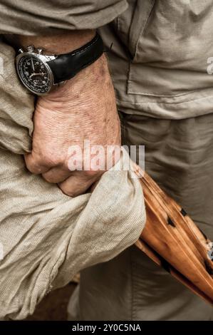 Ein Blick auf die rechte Hand eines älteren Herrn, der sich an einem geschlossenen Schirm und einem khakifarbenen Tuch festhält. Er ist in Khaki gekleidet Stockfoto