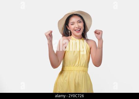 Fröhliche junge asiatische Frau in einem gelben Kleid, die ihre Hände in der Luft hochhält, mit einem glücklichen, erfolgreichen Ausdruck. Sie trägt einen Strohhut und Ohrringe Stockfoto