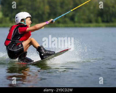 Junge oder Mädchen, sitzend auf Wakeboard, fröhlich, rote Schwimmweste, Wasserski und Sommerurlaub im Wakepark, Stras Pod Ralskem, Tschechische Republik, Europa Stockfoto