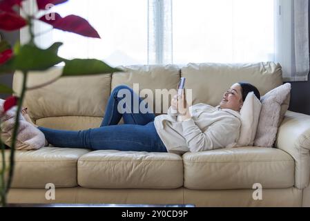 Junge Frau, die sich auf der Couch entspannt, im Wohnzimmer ihres Hauses auf ihrem Smartphone surft, einen ruhigen Moment genießt und digital verbunden ist Stockfoto