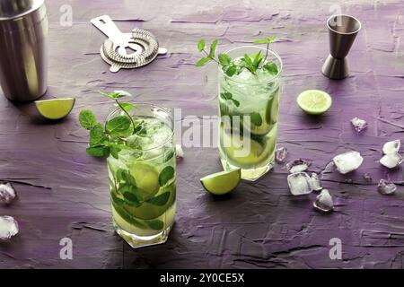 Mojito-Cocktail, zwei Gläser. Kaltes Sommergetränk mit Limette, frischer Minze und Eis. Kühles Getränk auf violettem Hintergrund mit Mischzubehör, Food Pho Stockfoto