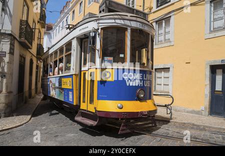 Eine gelb-blaue Straßenbahn schlängelt sich durch eine Kopfsteinpflasterstraße, die von historischen Gebäuden umgeben ist: Tram, Linie 28, Carros Electricos de Lisboa, Electricos de Stockfoto