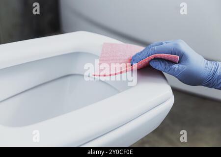 Frau Hand Nahaufnahme mit Gummischutzhandschuhen. Hausfrau, die Toilettenschüssel reinigt, Sitz mit Waschmittel, pinkfarbenes Tuch im Badezimmer oder auf öffentlichen Toiletten. Stockfoto