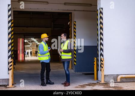 Porträt von zwei Arbeitern, die eine Pause von der Arbeit machen, Kaffee trinken, reden und sich ausruhen, auf der Laderampe stehen, im Freien. Kollegen trinken warmen Tee Stockfoto