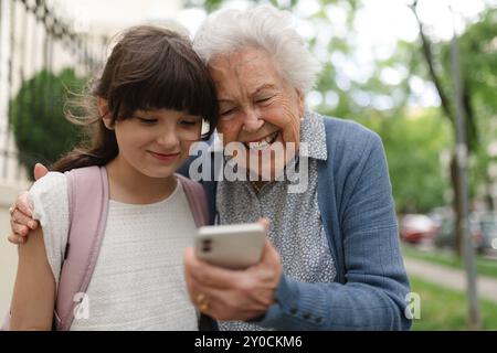 Oma holt das junge Mädchen von der Schule ab. Enkelin zeigt etwas auf dem Smartphone der Seniorengroßmutter. Stockfoto