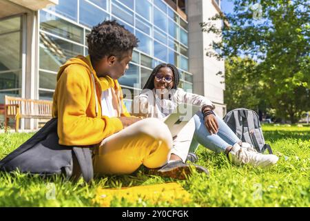 Seitenansicht von zwei afroamerikanischen Studenten, die einen Laptop benutzen, der auf dem Gras auf dem Campus sitzt Stockfoto