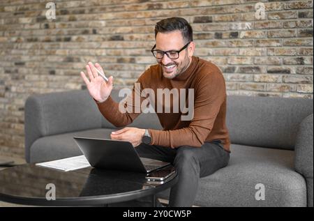 Fröhlicher junger Mann winkt Hand über Videoanrufe auf einem Laptop, während er an einem Online-Meeting im Büro teilnimmt Stockfoto