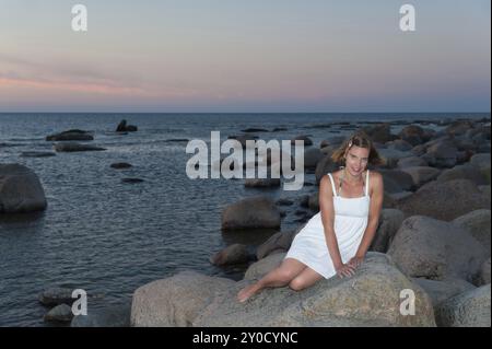 Attraktive junge Frau sitzt auf einem großen Felsen an der Ostseeküste. Das Bild erinnert an die kleine Meerjungfrau. Attraktive blonde Frau sitzt auf einem Stockfoto